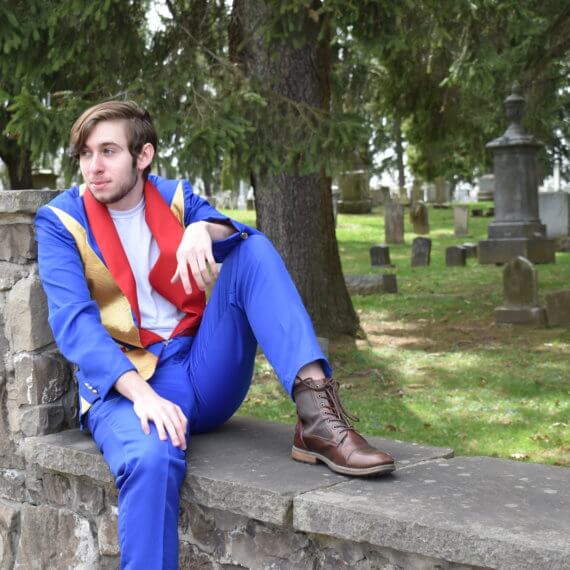A men’s sports jacket with royal blue sleeves, alternating royal blue and gold chevron pattern on the front and back of the torso, and red shawl lapels; paired with a white crew neck t-shirt, royal blue trousers, and brown man’s ankle boots. The model is sitting and leaning against a stone wall, in front of a nature filled cemetery.