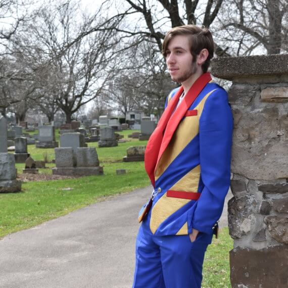 A men’s sports jacket with royal blue sleeves, alternating royal blue and gold chevron pattern on the front and back of the torso, and red shawl lapels; paired with a white crew neck t-shirt, and royal blue trousers. The model is leaning against a stone wall with a nature filled cemetery in the background