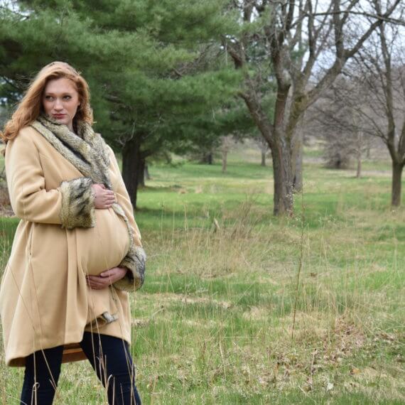 A tan colored maternity coat with accents of faux wolf fur on the collar, cuffs, and pocket lining. The model is holding the coat closed and protecting her belly while standing in a woodsy field