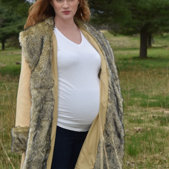 A tan colored maternity coat with accents of faux wolf fur on the collar, cuffs, and pocket lining. The coat is paired with a white maternity shirt and blue jeans. The model is standing in front of a woodsy background