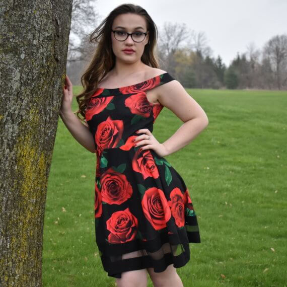 An above the knee, off the shoulder black dress with red roses printed on it. The model is leaning against a tree with a grass field in the background