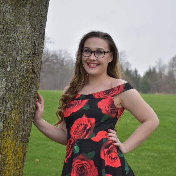 An above the knee, off the shoulder black dress with red roses printed on it. The model is resting a hand on a tree while smiling and looking into the distance, in front of an open field