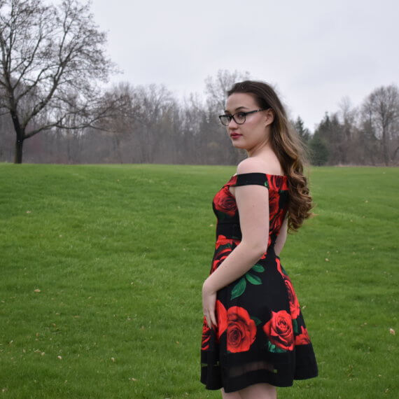 An above the knee, off the shoulder black dress with red roses printed on it. The model is turned away and looking over her shoulder at the camera while standing in an open field