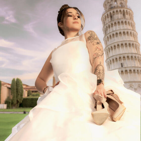 A sleeveless white organza wedding dress with a layered handkerchief hem. The model is holding a pair of nude high heels in front of the Leaning Tower of Pisa.