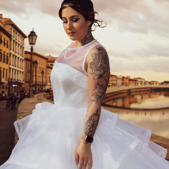 A sleeveless white organza wedding dress with a layered handkerchief hem. The model is standing in front of the Arno River in Pisa, Italy.