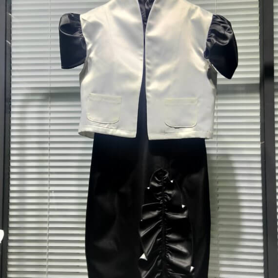 A two piece look of a black satin sleeveless shift dress with a mandarin collar and asymmetrical vertical ruffle on the skirt paired with a bolero length white satin jacket with contrasting black, set in, puff, cap short sleeves. The jacket has a stand up shawl collar