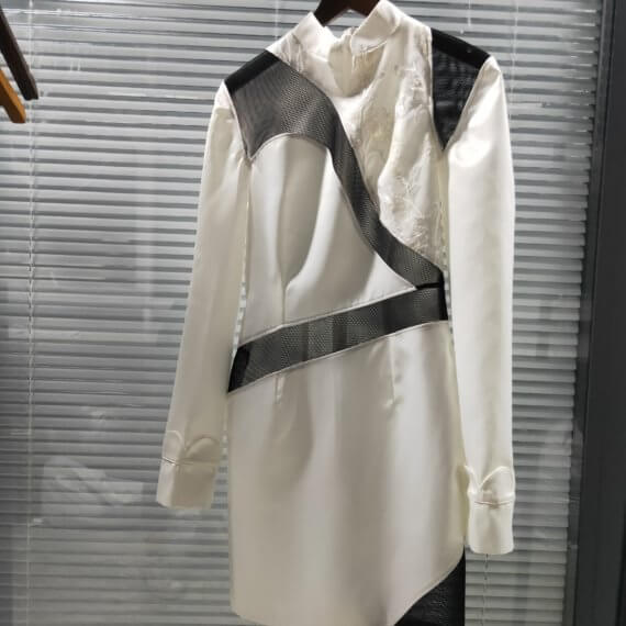 White satin mid thigh length long sleeved shift dress with a small v necked opening, banded collar and asymetrical black tulle insets at bottom left hem, left shoulder, and diagonal insets crossing the midsection and bodice. The upper left bodice has a a white floral embroidered overlay. The long sleeves feature a large scallope d piping.