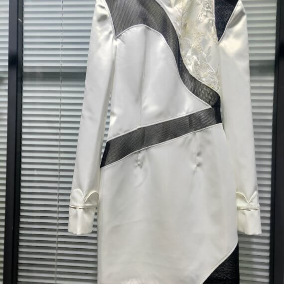 White satin mid thigh length long sleeved shift dress with a small v necked opening, banded collar and asymetrical black tulle insets at bottom left hem, left shoulder, and diagonal insets crossing the midsection and bodice. The upper left bodice has a a white floral embroidered overlay. The long sleeves feature a large scallope d piping.