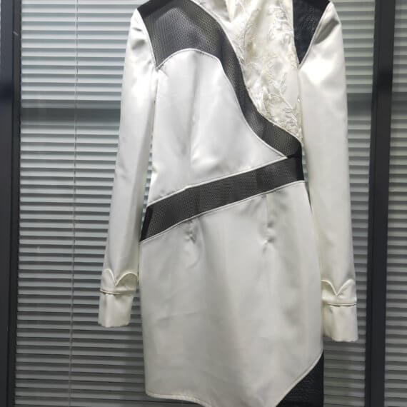 White satin mid thigh length long sleeved shift dress with a small v necked opening, banded collar and asymetrical black tulle insets at bottom left hem, left shoulder, and diagonal insets crossing the midsection and bodice. The upper left bodice has a a white floral embroidered overlay. The long sleeves feature a large scallope d piping.