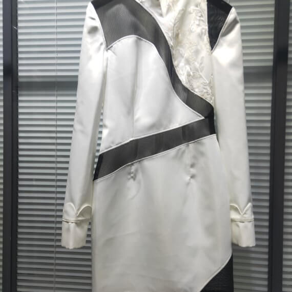 White satin mid thigh length long sleeved shift dress with a small v necked opening, banded collar and asymetrical black tulle insets at bottom left hem, left shoulder, and diagonal insets crossing the midsection and bodice. The upper left bodice has a a white floral embroidered overlay. The long sleeves feature a large scallope d piping.