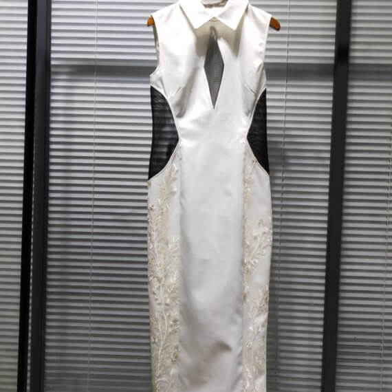 White satin tea length sleeveless shift dress with sheer black tulle elongated diamond keyhole. The dress has princess seams and a windsor collar. The sides of the dress include a white floral embroidered overlay and black tulle cut outs at the waist.