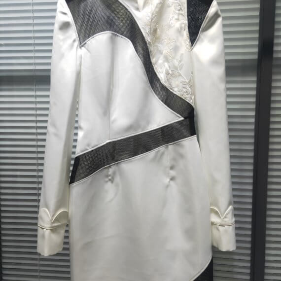 White satin mid thigh length long sleeved shift dress with a small v necked opening, banded collar and asymetrical black tulle insets at bottom left hem, left shoulder, and diagonal insets crossing the midsection and bodice. The upper left bodice has a a white floral embroidered overlay. The long sleeves feature a large scallope d piping.