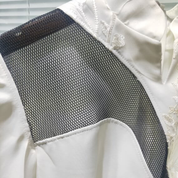 White satin mid thigh length long sleeved shift dress with a small v necked opening, banded collar and asymetrical black tulle insets at bottom left hem, left shoulder, and diagonal insets crossing the midsection and bodice. The upper left bodice has a a white floral embroidered overlay. The long sleeves feature a large scallope d piping.