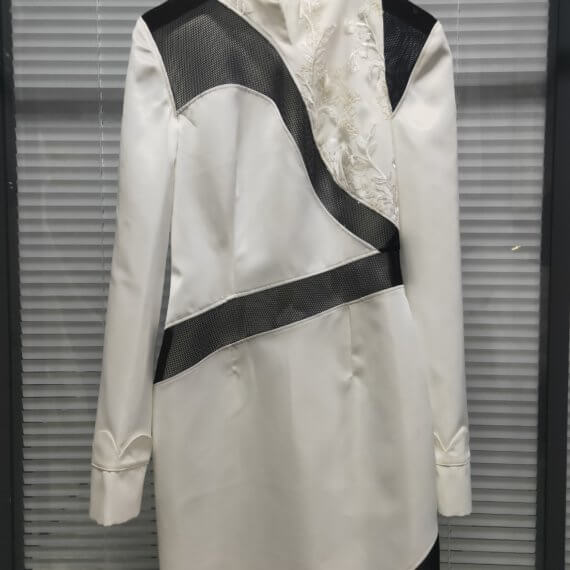 White satin mid thigh length long sleeved shift dress with a small v necked opening, banded collar and asymetrical black tulle insets at bottom left hem, left shoulder, and diagonal insets crossing the midsection and bodice. The upper left bodice has a a white floral embroidered overlay. The long sleeves feature a large scallope d piping.