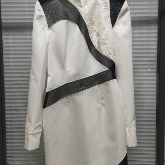 White satin mid thigh length long sleeved shift dress with a small v necked opening, banded collar and asymetrical black tulle insets at bottom left hem, left shoulder, and diagonal insets crossing the midsection and bodice. The upper left bodice has a a white floral embroidered overlay. The long sleeves feature a large scallope d piping.