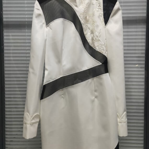 White satin mid thigh length long sleeved shift dress with a small v necked opening, banded collar and asymetrical black tulle insets at bottom left hem, left shoulder, and diagonal insets crossing the midsection and bodice. The upper left bodice has a a white floral embroidered overlay. The long sleeves feature a large scallope d piping.
