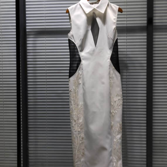 White satin tea length sleeveless shift dress with sheer black tulle elongated diamond keyhole. The dress has princess seams and a windsor collar. The sides of the dress include a white floral embroidered overlay and black tulle cut outs at the waist.