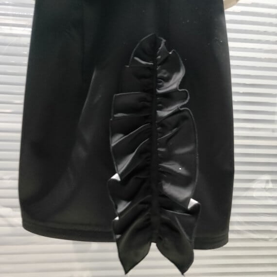 A two piece look of a black satin sleeveless shift dress with a mandarin collar and asymmetrical vertical ruffle on the skirt paired with a bolero length white satin jacket with contrasting black, set in, puff, cap short sleeves. The jacket has a stand up shawl collar.