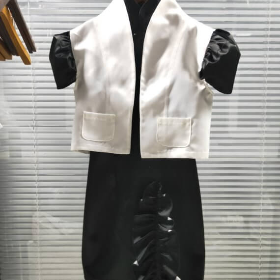 A two piece look of a black satin sleeveless shift dress with a mandarin collar and asymmetrical vertical ruffle on the skirt paired with a bolero length white satin jacket with contrasting black, set in, puff, cap short sleeves. The jacket has a stand up shawl collar
