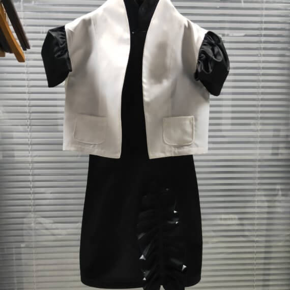 A two piece look of a black satin sleeveless shift dress with a mandarin collar and asymmetrical vertical ruffle on the skirt paired with a bolero length white satin jacket with contrasting black, set in, puff, cap short sleeves. The jacket has a stand up shawl collar