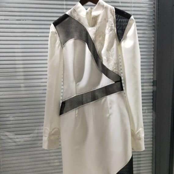 White satin mid thigh length long sleeved shift dress with a small v necked opening, banded collar and asymetrical black tulle insets at bottom left hem, left shoulder, and diagonal insets crossing the midsection and bodice. The upper left bodice has a a white floral embroidered overlay. The long sleeves feature a large scallope d piping.